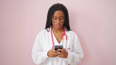 Afro-Amerikalı kadın doktor, pembe arka plan üzerinde akıllı telefon kullanarak gülümsüyor.