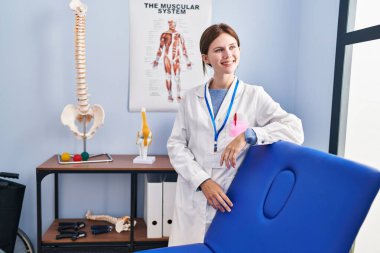 Genç sarışın kadın fizyoterapist gülümsüyor rehabilitasyon merkezindeki masaj masasına yaslanıyor.