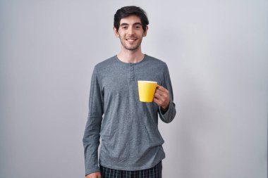 Genç İspanyol adam pijama giyip yüzünde mutlu ve havalı bir gülümsemeyle bir fincan kahve içiyor. şanslı kişi. 