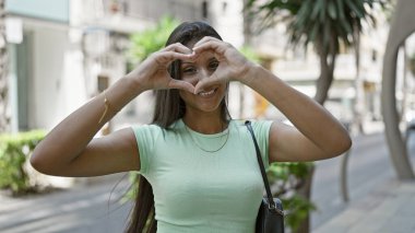 Genç Latin kadın gülümsüyor. Kendine güveniyor. Sokakta elleriyle kalp hareketi yapıyor.