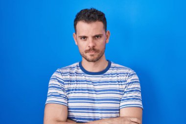 Young hispanic man standing over blue background skeptic and nervous, disapproving expression on face with crossed arms. negative person. 