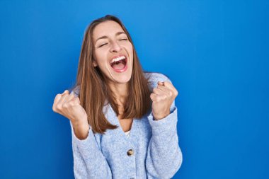 Young woman standing over blue background celebrating surprised and amazed for success with arms raised and eyes closed. winner concept. 