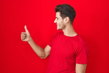 Young hispanic man standing over red background looking proud, smiling doing thumbs up gesture to the side 