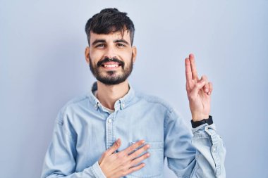 Mavi arka planda sakallı genç bir İspanyol adam gülümsüyor, elleri göğsünde ve parmakları havada küfür ediyor, sadakat yemini ediyor. 