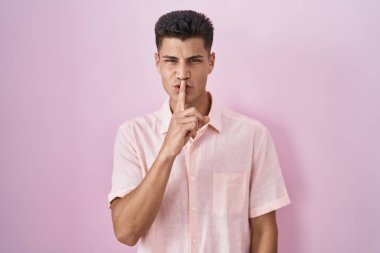 Pembe arka planda dikilen genç İspanyol adam parmakları dudaklarında sessiz olmak istiyor. sessizlik ve gizli konsept. 