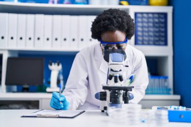 Young african american woman scientist writing on document using microscope at laboratory