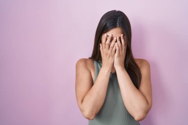 Hispanic woman standing over pink background with sad expression covering face with hands while crying. depression concept. 