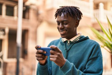 Afro-Amerikalı adam gülümsüyor. Sokakta akıllı telefondan videolar izliyor.