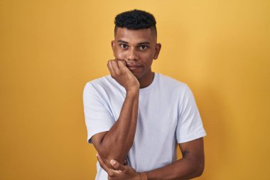 Young hispanic man standing over yellow background looking stressed and nervous with hands on mouth biting nails. anxiety problem. 