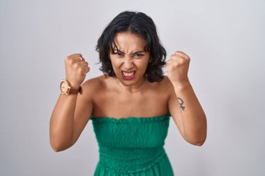 Young hispanic woman standing over isolated background angry and mad raising fists frustrated and furious while shouting with anger. rage and aggressive concept. 
