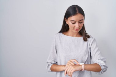 Beyaz arka planda duran genç İspanyol kadın kol saatindeki saati kontrol ediyor, rahat ve kendinden emin. 