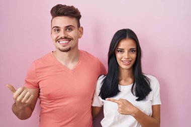 Pembe arka planda duran genç İspanyol çift arka tarafı işaret ediyor. Elleri ve başparmakları havada, kendine güvenen. 