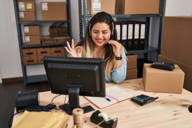 Young hispanic woman working at small business ecommerce wearing headset doing ok sign with fingers, smiling friendly gesturing excellent symbol 