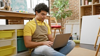 Sanat stüdyosunda dizüstü bilgisayar kullanarak gülümseyen genç Latin sanatçı.