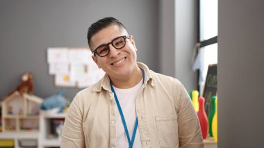 Young hispanic man preschool teacher smiling confident standing at kindergarten