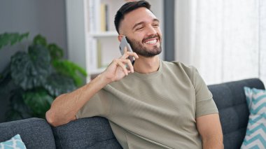 Genç İspanyol adam telefonda konuşuyor. Evdeki kanepede oturuyor.