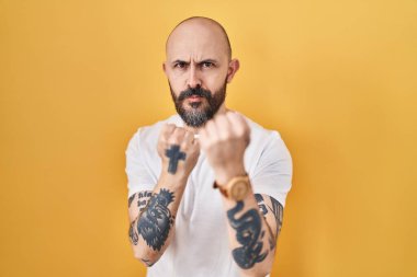 Sarı arka planda dövmeleri olan genç İspanyol bir adam. Yumruk savunma hareketiyle dövüşmeye hazır. Kızgın ve üzgün bir yüz, sorundan korkuyor. 