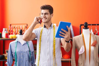 Young hispanic man tailor talking on smartphone using touchpad at atelier