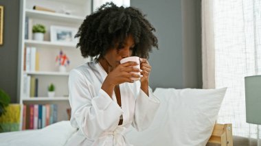 Afro-Amerikalı kadın yatakta oturmuş, yatak odasında kahve içiyor.