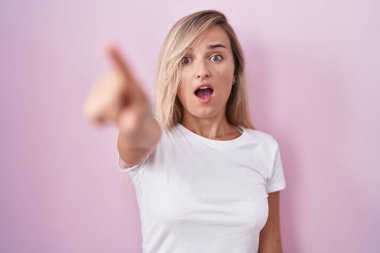 Young blonde woman standing over pink background pointing with finger surprised ahead, open mouth amazed expression, something on the front 