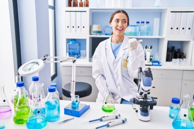 Genç esmer kadın, bilim adamlarının laboratuarında çalışıyor. Gülümsüyor, gülümsüyor ve başparmağıyla yan tarafa bakıyor.. 