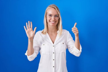 Young caucasian woman standing over blue background showing and pointing up with fingers number six while smiling confident and happy. 