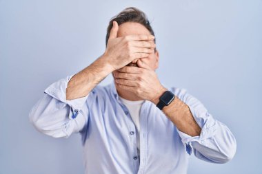 Orta yaşlı beyaz bir adam mavi arka planda duruyor gözlerini ve ağzını elleriyle kapatıyor, şaşırmış ve şok olmuş. duygu gizleme 