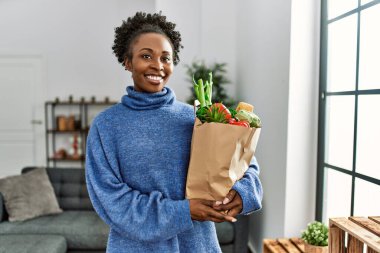 Afro-Amerikalı bir kadın, elinde alışveriş poşeti tutarak gülümsüyor.