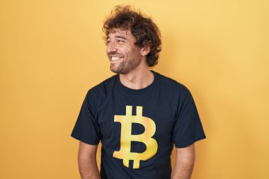 Bitcoin tişörtü giyen İspanyol genç adam yüzünde gülümsemeyle yan yana bakıyor, doğal bir ifade. Kendinden emin gülen. 