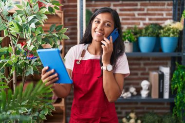 Young beautiful hispanic woman florist talking on smartphone using touchpad at flower shop