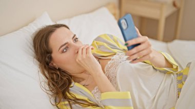 Yatak odasında uzanmış akıllı telefon kullanan genç sarışın kadın.