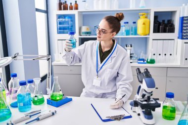 Young woman scientist writing on clipboard holding test tube at laboratory