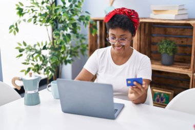 Evdeki koltukta oturan Afrikalı Amerikalı kadın dizüstü bilgisayar ve kredi kartı kullanıyor.