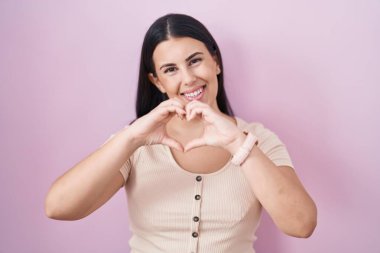 Young hispanic woman standing over pink background smiling in love doing heart symbol shape with hands. romantic concept. 