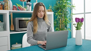 Genç beyaz kadın yemek odasında masada dizüstü bilgisayar kullanıyor.