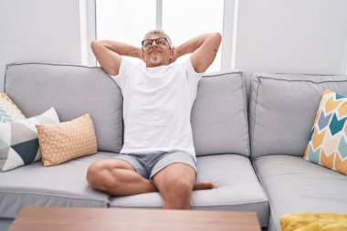 Orta yaşlı, kır saçlı, elleri başının üstünde, evinde kanepede oturan bir adam.
