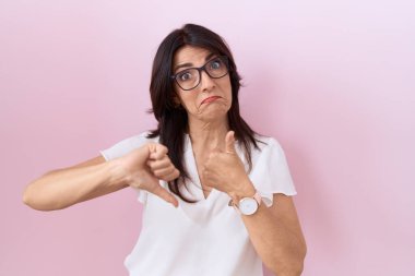Orta yaşlı İspanyol kadın, günlük beyaz tişört ve gözlük giyip baş parmak hareketi yapıyor, anlaşmazlık ve anlaşma ifadesi. çılgın çatışma 
