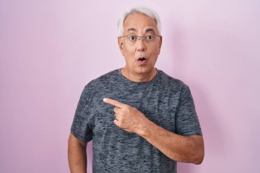 Pembe arka planda duran gri saçlı orta yaşlı bir adam parmaklarını yana çevirerek şaşırdı, ağzı açık hayret içinde kaldı.. 