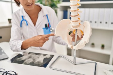Young beautiful hispanic woman doctor pointing with finger to anatomical model of vertebral column at clinic