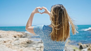 Genç sarışın kadın deniz kenarında kalp hareketi yapıyor.