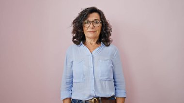Middle age hispanic woman standing with serious expression over isolated pink background