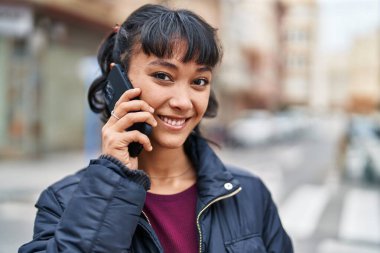 Genç, güzel, İspanyol bir kadın gülümsüyor ve caddede akıllı telefondan konuşuyor.