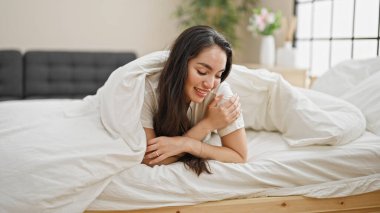 Young beautiful hispanic woman lying on bed covering with bedsheet at bedroom