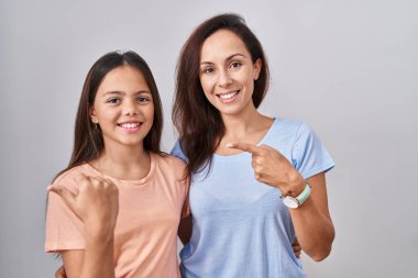 Genç anne ve kızı beyaz arka planda dikiliyorlar. Elleri ve başparmakları havada, kendinden emin gülümsüyorlar. 