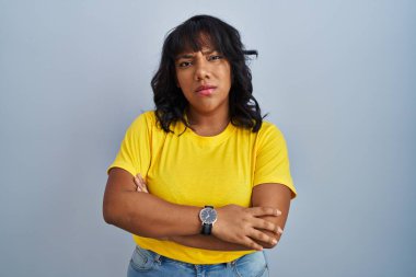 Mavi arka planda duran İspanyol kadın şüpheci ve gergin, yüzünde çapraz kollu tasvip etmeyen ifade. negatif kişi. 