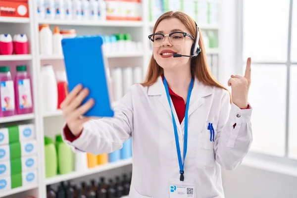 在药店里工作的红头发女人做着视频通话 带着平板电脑微笑 手指手画脚地指向旁边 — 图库照片