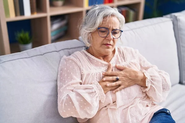Middle age grey-haired woman sitting on sofa suffering heart attack at home