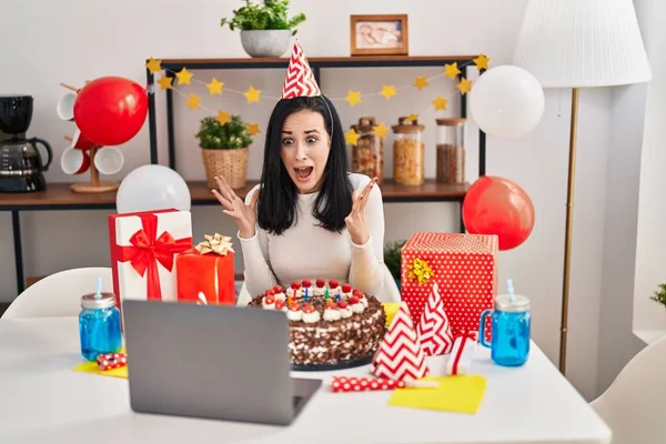 Hispanic woman celebrating birthday with big chocolate cake doing video call celebrating victory with happy smile and winner expression with raised hands