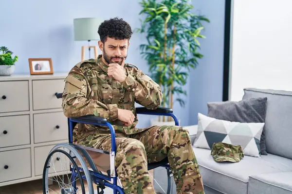 Arab man wearing camouflage army uniform sitting on wheelchair looking confident at the camera with smile with crossed arms and hand raised on chin. thinking positive.