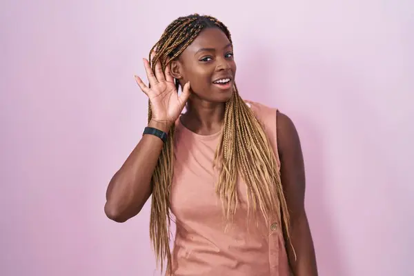 一个有着辫子的非裔美国女人站在粉红的背景上微笑着 两手空空地听着谣言或闲话 聋的概念 — 图库照片